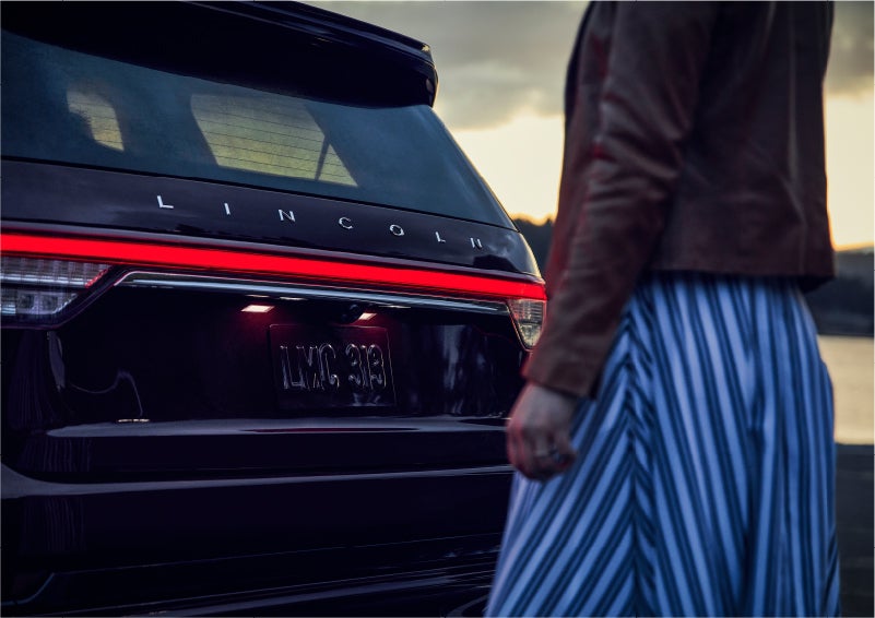A person is shown near the rear of a 2023 Lincoln Aviator® SUV as the Lincoln Embrace illuminates the rear lights | Hooks Lincoln in Fort Worth TX