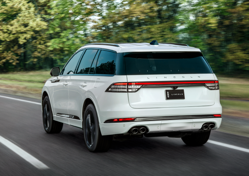 A white 2025 Lincoln Aviator® SUV is being driven away down a road lined with colorful trees. | Hooks Lincoln in Fort Worth TX