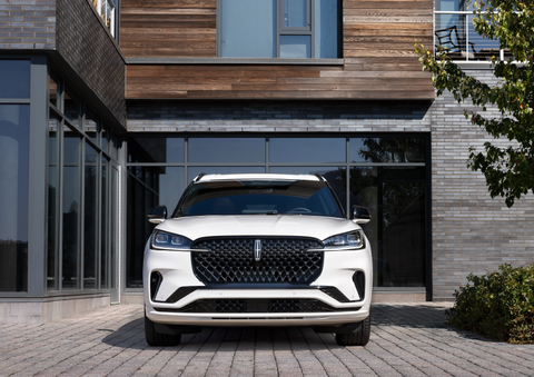 A white 2025 Lincoln Aviator® SUV is shown parked outside a modern building with gray brick and wood accents. | Hooks Lincoln in Fort Worth TX