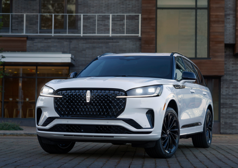 A white 2025 Lincoln Aviator® SUV is shown parked outside a modern building with gray brick and wood accents. | Hooks Lincoln in Fort Worth TX