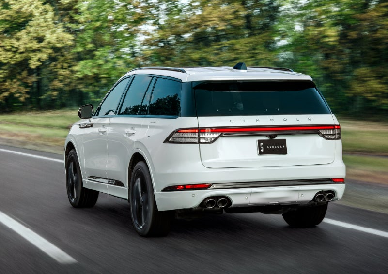 A white 2026 Lincoln Aviator® SUV is being driven away down a road lined with colorful trees. | Hooks Lincoln in Fort Worth TX